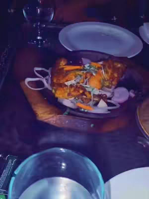 The image is a tabletop shot capturing a plate of food, likely a meal, on a dark, possibly wooden surface. Surrounding elements include a partially visible glass of liquid, potentially water or a beverage, positioned closely to the center-left of the frame. The food itself is the highlight, with a mix of golden-brown components, possibly fried or grilled, alongside fresh vegetables such as carrots and sliced onions, garnished with some green garnish. The shot is angled to provide a top-down perspective, and the colors are warm.