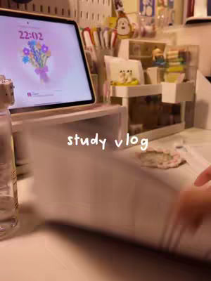 A person's desk setup is shown with a laptop displaying a floral image on its screen. In front of the laptop, a book is being held up, featuring the overlay text in the middle. The surroundings include pens, decorative items, and a bottle, suggesting a study environment. The color palette is soft, with neutral tones.