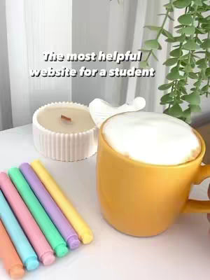 The image shows a close-up of a yellow mug filled with a foamy drink, likely a latte or cappuccino, on a white surface. Next to the mug is a white candle in a ribbed container, and a set of colorful markers. In the background, there's a glimpse of leaves and a white curtain.