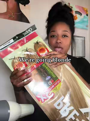 A woman with dark hair holds up a package of blonde hair extensions, looking towards the camera with an open mouth expression. The text overlay reads, "We're going blonde."