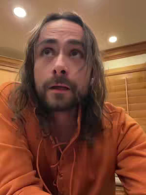 A man with long brown hair and a beard is looking upward with an open mouth, dressed in an orange shirt, in a room with a light fixture and wooden blinds visible.