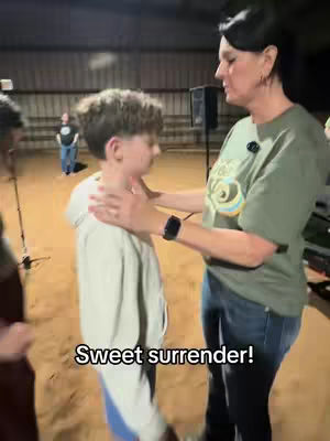 A woman in a green t-shirt and jeans stands to the right, holding a young boy around the shoulders and neck. The boy is facing away from the camera with his eyes closed. Both are standing in an open area. Text overlayed on the image says "Sweet surrender!".