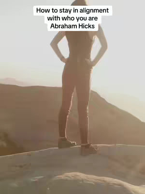 A woman stands on a rock, seen from the back, with arms at her sides and the horizon in the background. The sun is low in the sky, creating a warm tone and highlighting the silhouette of the figure and the mountain range.
