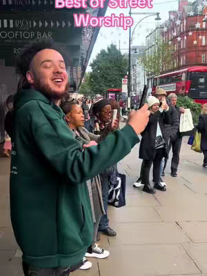 A man in a green hoodie is holding a phone and filming on a busy street with people around him. The image is brightly lit with a focus on the central figure and surrounding pedestrians, and a red bus can be spotted in the background.