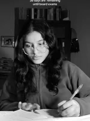 A grayscale image shows a person, likely a student, studying. The person has long, wavy hair and wears glasses. They are sitting at a desk, writing on a piece of paper. There is text overlayed at the top: "36 days are remaining until board exams". The background is slightly blurred, suggesting a room with a bookshelf.