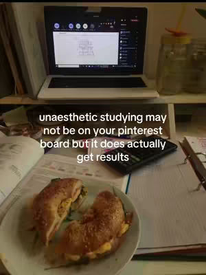 A photo of a desk setup. A laptop is open in the background. In the foreground, there's a plate with a sandwich and some papers.