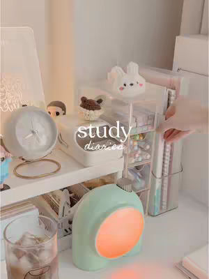 The cover image showcases a cozy desk setup with various items. A mint green lamp with an orange glow sits at the bottom center. To the left is a clear glass with a drink in it. In the background, there's a shelf with a gray clock and some decorative items including a small plush. In the center is a clear multi-tiered storage shelf filled with stationary. The words 'study diaries' are written in a script font. The overall composition is soft and aesthetically pleasing, with a focus on creating a workspace.