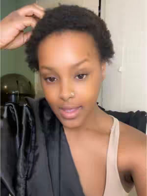 The cover image features a close-up of a person's face, likely the person recording the video. The person has short, dark, and curly hair. They are looking at the camera with a slight smile and their right hand is touching their hair. They are wearing a light-colored tank top and a black robe or jacket is draped over their shoulders. The background is indistinct, with a glimpse of what seems to be a piece of black equipment. The lighting appears to be neutral and the composition focuses on the person's face, creating an intimate and personal feel.