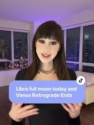 A woman with long brown hair, parted in the middle with bangs, is centered in the frame. She is wearing a black top and a necklace. The background shows an apartment with large windows overlooking a cityscape. A light blue text overlay is positioned at the lower mid-section of the image.