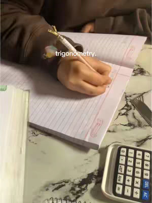 A person is writing with a pen in a notebook in which the word trigonometry is written. The picture is taken from above and includes a calculator and a notebook.