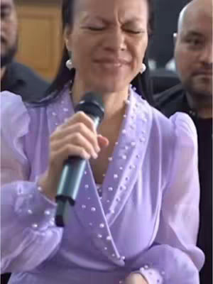 A woman with her eyes closed, holding a microphone. She wears a purple, pearl-embellished outfit. Two men are blurred in the background.