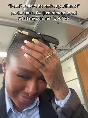 A person with short dark hair is seen close-up with their hand to their forehead. They have a nose piercing and are wearing sunglasses on top of their head. The background has some metallic structures.