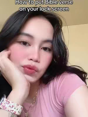 A light-skinned woman with long, dark hair poses for a selfie. She is wearing a pink t-shirt and a white watch with a floral pattern on her wrist. She is holding her hand near her face, resting her chin on her fingers. Her lips are colored with pink lipstick. The background is a light beige color.