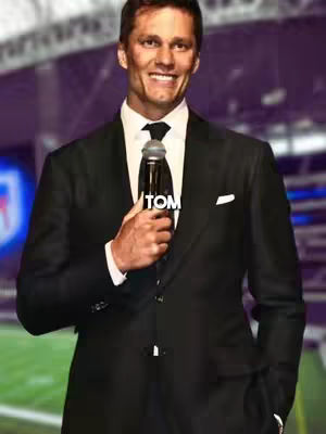 Tom Brady in a black suit holding a microphone and smiling. The background is blurred featuring a stadium setting with a purple-ish hue.