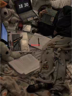 The image shows a person lying in bed, surrounded by study materials. A laptop displaying 18:30 sits open on the top, and papers and books are scattered around. The person appears to be resting with a textbook covering their face, and wearing Snoopy patterned pajama pants. The overall scene suggests a study session.