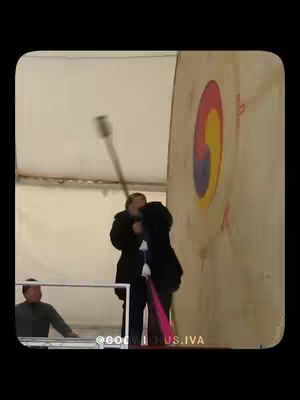 A person in a black outfit swings a mallet at a large target with a Korean symbol. Another person is behind them.