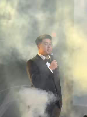 A man in a dark suit and bow tie stands in a room with smoke or fog, holding a microphone. The lighting is dramatic and there is a cloud of gray smoke surrounding him.