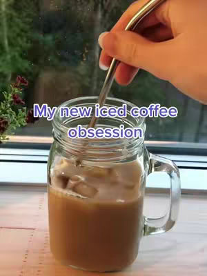 A hand holding a metal stirring stick is mixing coffee in a clear glass mug with ice. The mug is mostly filled with a light brown liquid, and it sits in front of a window with green foliage visible outside.