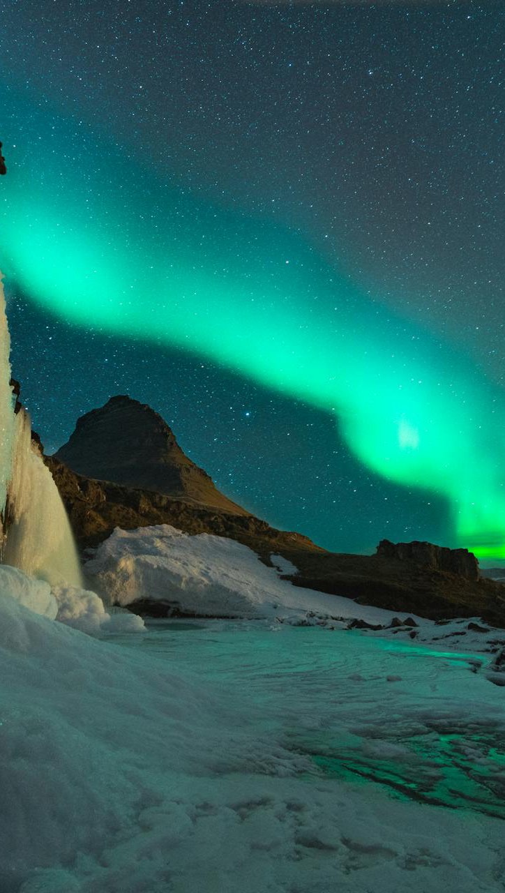 A scenic night shot of a mountain landscape under the vibrant green glow of the aurora borealis. A waterfall cascades down a cliff in the left foreground, with snow covering the lower areas. The sky is filled with stars and the bright green light of the aurora.