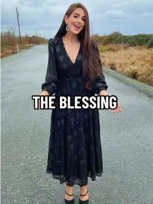 A woman in a black dress stands on a wet road, with buildings in the background. Text overlayed on the image reads "THE BLESSING".