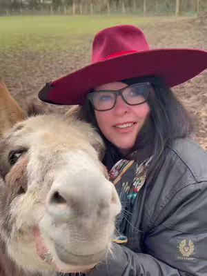 The cover image features a woman hugging a donkey. She is wearing a burgundy hat, glasses, and a black coat. The donkey is positioned to the left of the frame, with its head close to the woman's, creating a warm, tactile feel. The background appears to be an outdoor setting, likely a field, with muted tones. The overall composition is a blend of warmth and natural interaction.