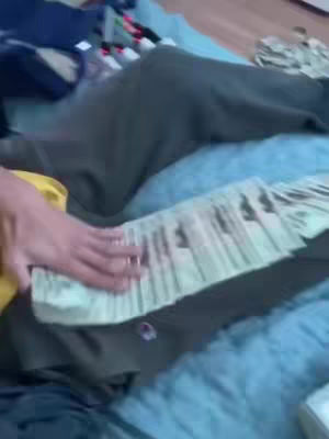The cover image is slightly blurry and depicts a person's hand handling a large stack of US currency, primarily twenty-dollar bills. The hand is positioned over a dark green garment, likely a sweatshirt, which is resting on a blue-grey surface, possibly a bed or couch. There are additional stacks of money visible in the background. The lighting appears to be natural, with a warm tone, however the image lacks sharp focus.