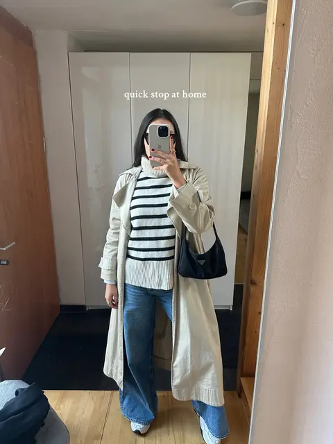 A woman stands in a room, facing a mirror and taking a selfie with her phone. She is wearing a striped sweater, jeans, a trench coat, and white sneakers. A small black purse hangs on her shoulder. The background includes a white cabinet and a wooden door frame