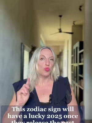 A woman with long blonde hair and a dark top is talking directly to the camera, pointing with her index finger. The background shows a hallway with a ceiling fan and framed artwork.