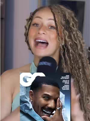 A woman with blond and brown braided hair is holding a microphone and smiling. She has a magazine partially covering her body showing a black man on the cover of the magazine. The background is blurry and shows what looks like a light-colored wall.