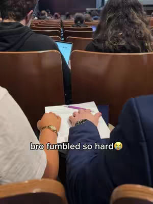 The image shows a classroom setting with rows of wooden chairs. The focus is on two sets of hands, one with a watch and the other holding a pen over a paper. Text overlay at the bottom reads 'bro fumbled so hard' with an emoji.
