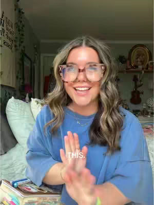 A smiling woman with glasses is shown indoors. She's wearing a blue shirt and her hands are up in a prayer-like gesture, with the word 'THIS' in front. The background includes a window, plants, and a room interior.