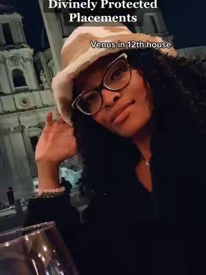 A woman wearing glasses and a hat is the focal point of this low-angle shot. The background features a well-lit building at nighttime, with vertical structures and arched windows. The image is warm-toned with overall good lighting.