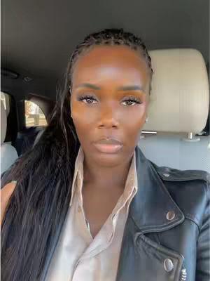 A medium shot shows a woman seated in the driver's seat of a car, looking directly at the camera with a serious expression. She has dark hair, styled in braids, and is wearing a black leather jacket over a light-colored top. The background suggests the interior of a car with a visible headrest.