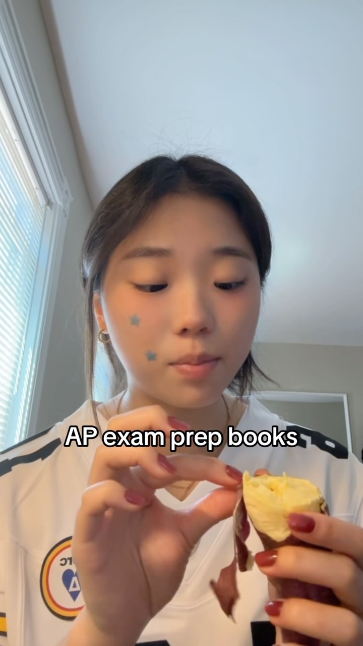 A young woman is holding a piece of a sweet potato, with blue star stickers on her cheeks. The background is a plain white wall with a window.