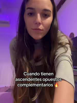 A woman with long brown hair looks directly at the camera. Text in white overlayed at the bottom of the video reads, 'Cuando tienen ascendentes opuestos complementarios' with a fire emoji. The background is a soft, purple-toned room.