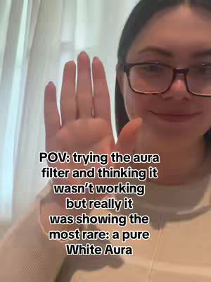 A selfie-style shot featuring a young woman with dark hair and glasses. She is holding her hand up, palm towards the camera, with her fingers slightly spread. The background is soft, and it appears to be an indoor setting. Text overlayed on the image includes 'POV: trying the aura filter and thinking it wasn't working but really it was showing the most rare: a pure White Aura'.