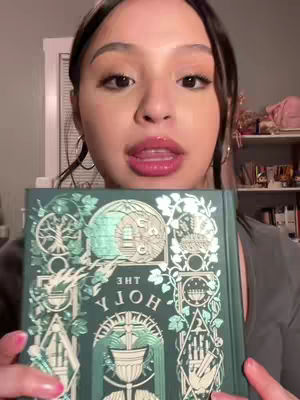 A woman with brown hair and makeup is holding a green book with gold and teal decorative designs on the cover. She is looking towards the camera with her mouth open as if she is speaking. The background is blurry.