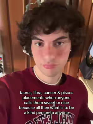 A selfie-style video featuring a young man with curly brown hair, wearing a maroon sweater and a pearl necklace, set against a room with wood-paneled walls. On the background, a text overlay indicates that the video may be an astrology content. The man looks directly at the camera with a neutral expression.