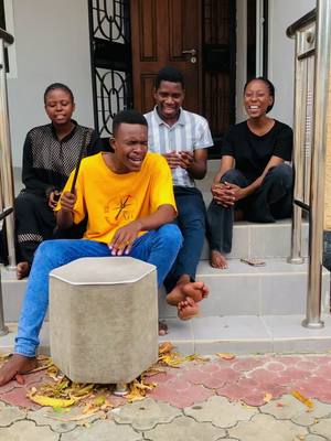 A color photo shows four young individuals, three males and one female, seated on a set of white steps outside a building. The main focus is on the young man in a yellow shirt sitting on a circular stool, appearing to sing with passion. Other individuals seated on the stairs show a mix of expressions, including smiles and engagement. The lighting is bright, and the overall composition is candid, suggesting a moment of musical performance or worship.