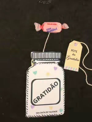 A still life composition against a dark background. A large, hand-drawn jar shape labeled 'GRATIDÃO' (gratitude) is centered. Attached is a pink candy labeled 'Yogurte 100', connected to the jar with a string. To the right, a tag with 'POTE da Gratidao' on it.