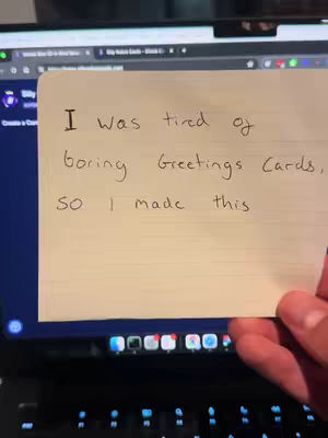 A hand holds up a cream-colored note. The note is handwritten with black ink, containing the text 'I was tired of boring Greetings Cards, so I made this.' In the background, a laptop is visible, its screen displaying a website. The composition is a medium shot, with the focus on the note and the text.