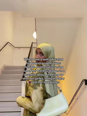 A woman wearing a green hijab and abaya is standing on a staircase, with a staircase and a light in the background, and a white handbag hanging on her shoulder. White text is overlayed on the image.