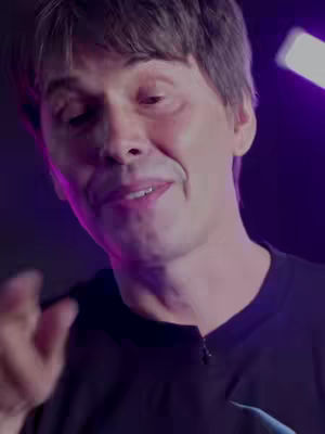 A medium shot of a man with short brown hair and a light complexion facing towards the camera. The man is wearing a black shirt. The background is a blurred mix of purple and dark tones, with a lighter purple light effect on the right. He appears to be talking with his mouth open slightly, and his left hand is raised near his chest. The overall lighting is moderate, and the focus is primarily on the man's face and upper body.