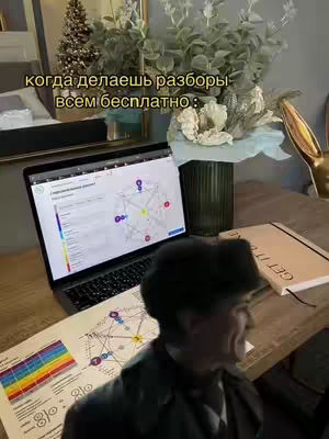 A person is seated behind a wooden desk. On the desk is a laptop displaying a complex diagram, a piece of paper with a similar diagram and color key, a notebook, and other decorative items. The background shows part of a bed, a lit Christmas tree, and a vase with flowers.