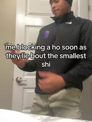 A man stands indoors, slightly angled to the left, holding a phone and looking towards the right. He is wearing a black zip-up jacket over a blue shirt and beige pants. The background includes a white door. The overlaid text reads, 'me blocking a ho soon as they lie bout the smallest shi.'