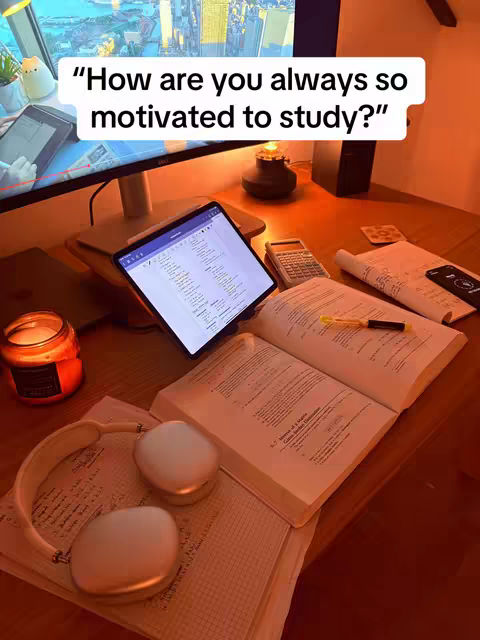 A well-lit desk setup with a computer monitor displaying a cityscape, a tablet, an open book, headphones, and a lit candle. The image has a warm, inviting feel, suggesting a study or workspace.