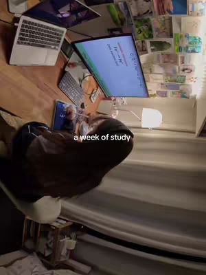 A fisheye view captures a person working at a desk covered in various items: a laptop, a large monitor displaying text, a keyboard, a tablet, and stacks of papers. Behind the person, there are drawings and sticky notes affixed to the wall with a soft light from a bedside lamp.