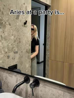 A woman with blonde hair stands partially visible in a doorway, wearing sunglasses and a black top. The setting appears to be a modern bathroom with a marble-like textured wall in view. The mirror reflects the bathroom fixtures, emphasizing a modern aesthetic.