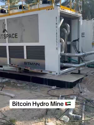 The image shows a large, white industrial machine housed inside a shipping container, labeled with the Bitmain logo. The container is open, revealing pipes and machinery. Text at the bottom reads 'Bitcoin Hydro Mine' and a UAE flag. The ground is covered with sand and some cables.
