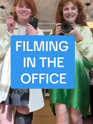 Two women are standing close together, both smiling at the camera. A large, blue rectangle in the center of the image has white text reading "FILMING IN THE OFFICE". The woman on the left is holding a phone and a small black object, and is wearing a cream-colored jacket. The woman on the right is holding a camera, and is wearing a green dress.